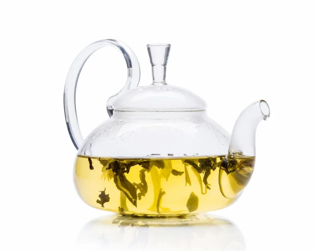 Glass teapot containing clear yellow tea with visible tea leaves, resting on a white surface.