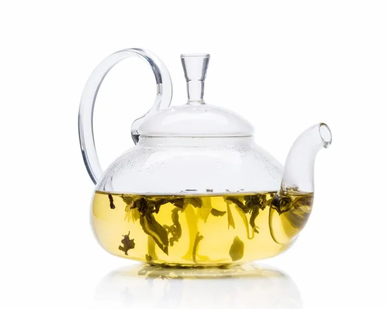 Glass teapot containing clear yellow tea with visible tea leaves, resting on a white surface.