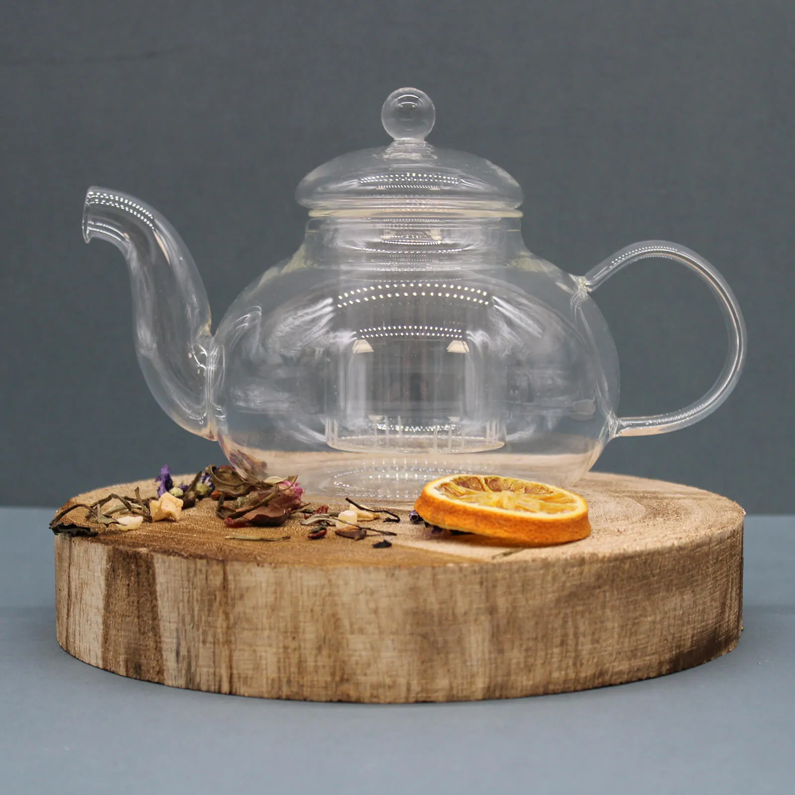 A clear glass teapot sits on a round wooden board with dried tea leaves and a slice of dried orange beside it, against a plain gray background.