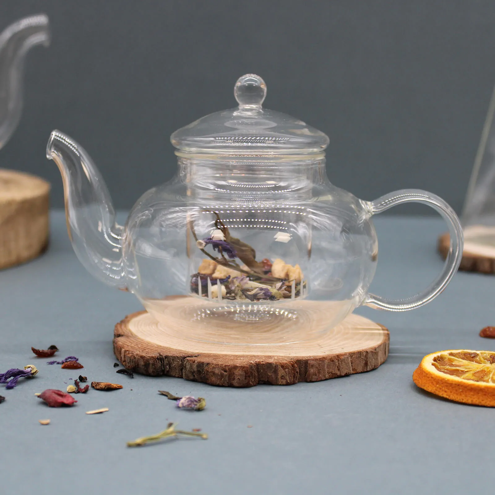 A clear glass teapot sits on a wooden coaster, containing loose dried herbs and flowers. Dried fruit and scattered tea leaves are nearby on a gray surface.