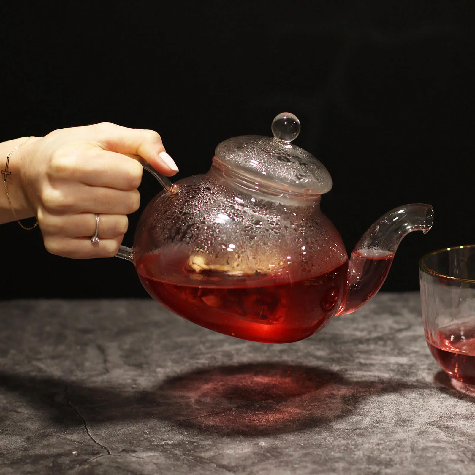 A hand pours red tea from a glass teapot into a glass cup on a dark surface, with steam visible on the teapot.