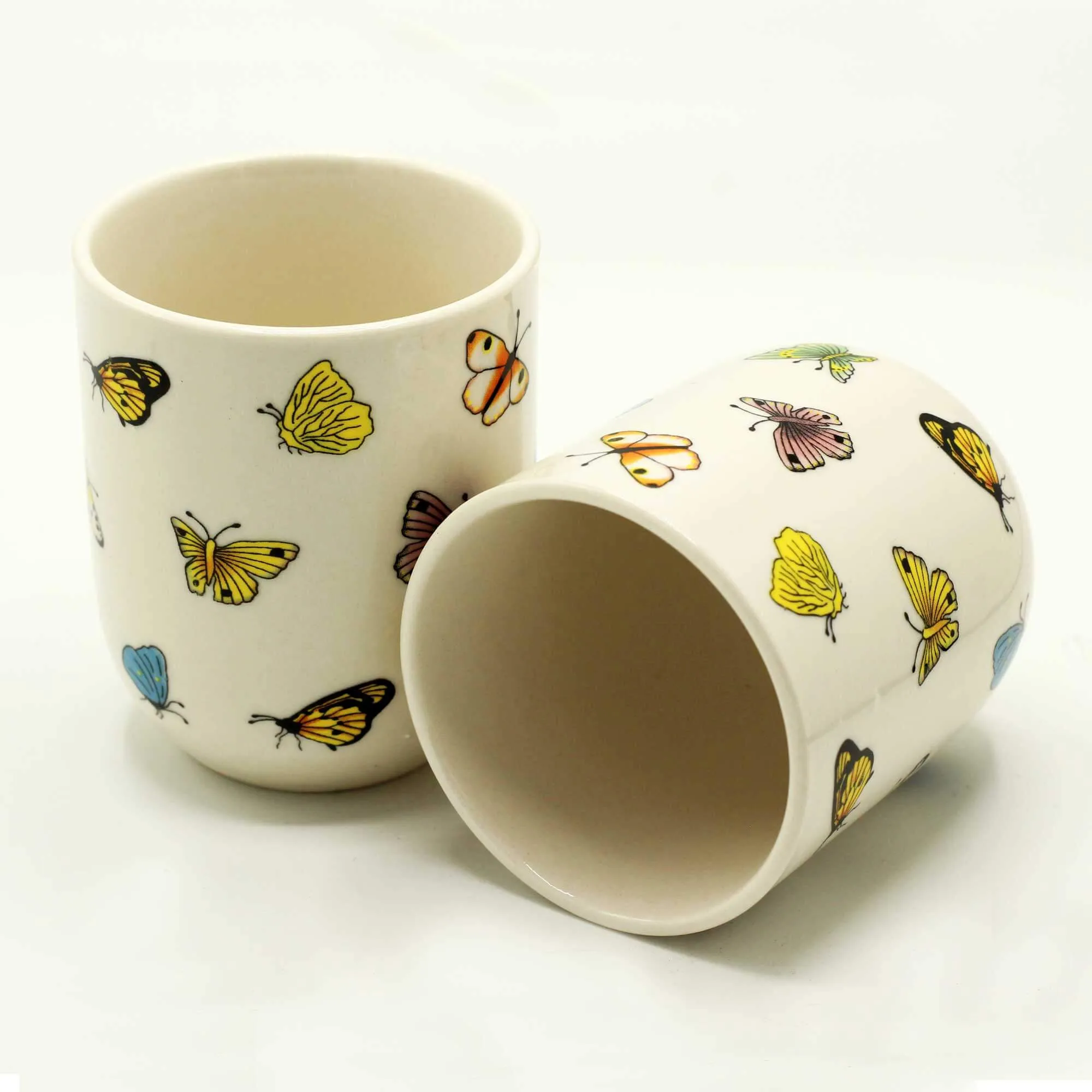 Two white ceramic cups with colorful butterfly and leaf patterns; one cup stands upright while the other lies on its side.