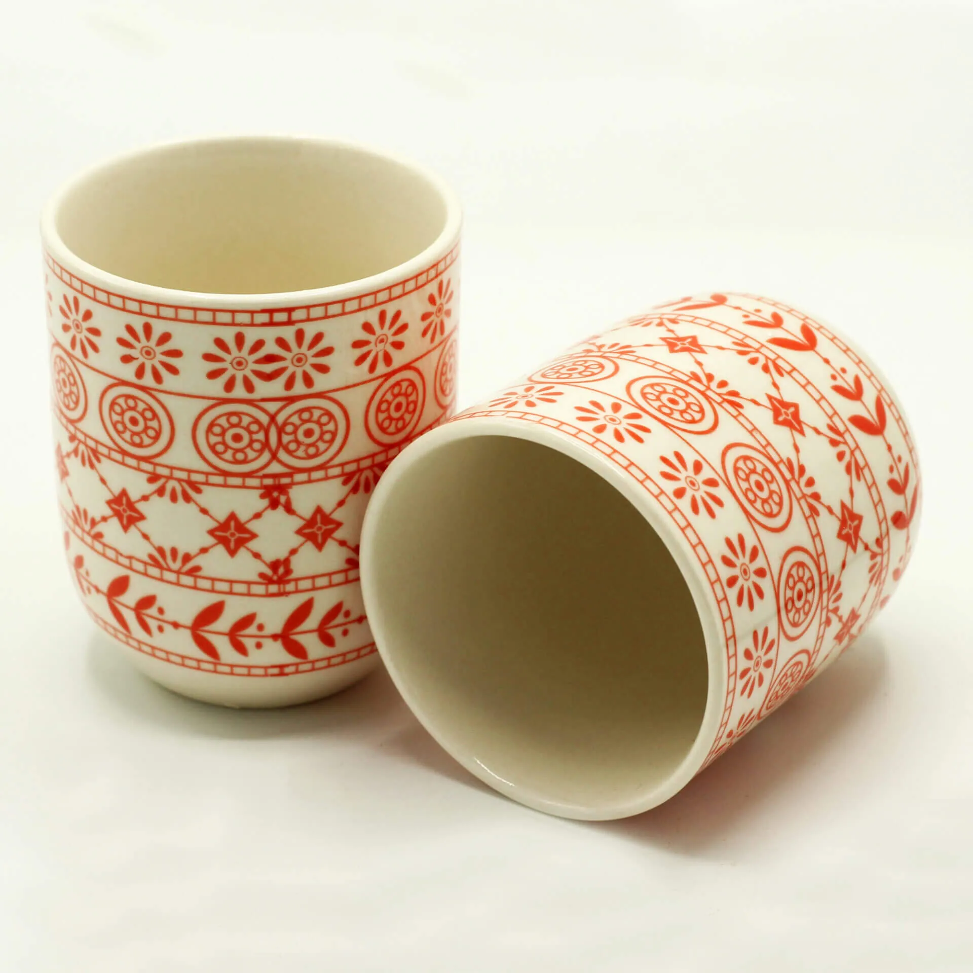 Two white ceramic cups with red floral and geometric patterns; one standing upright and the other lying on its side on a white background.