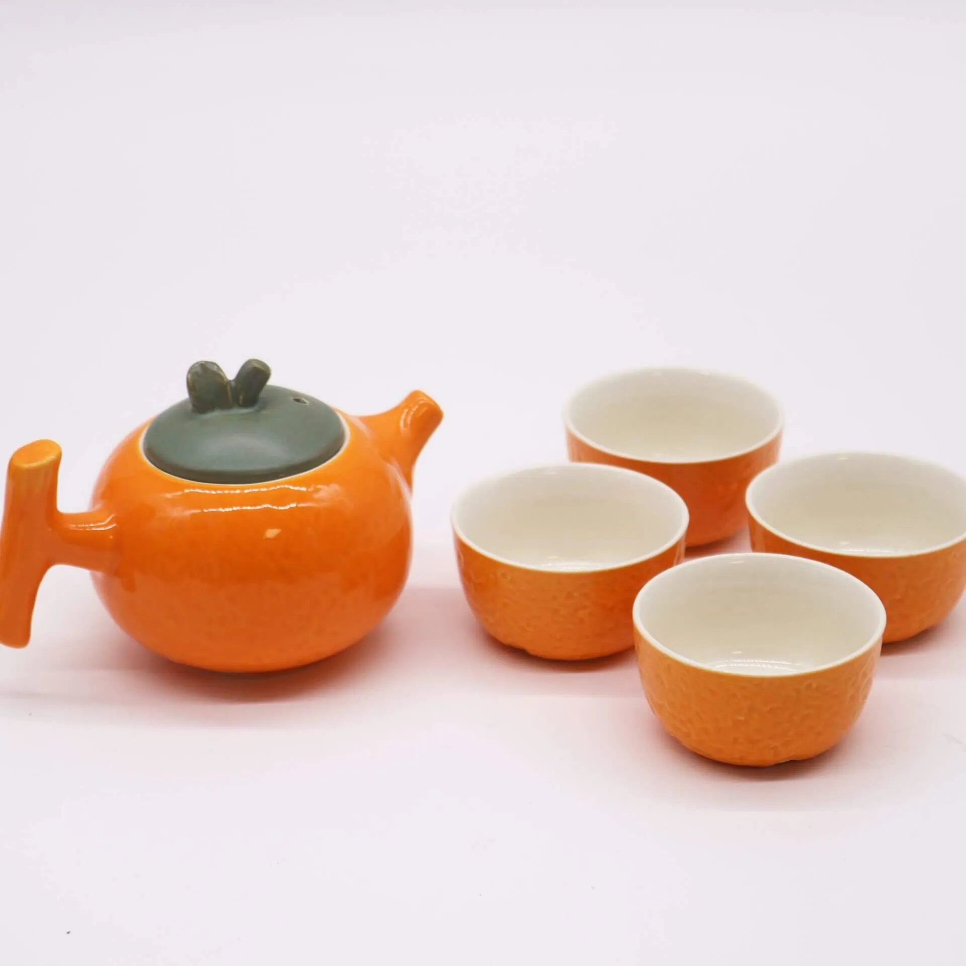 An orange teapot with a green lid is accompanied by four matching orange teacups, all placed on a white background.