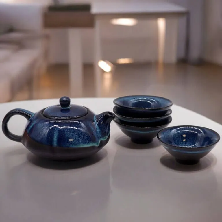 A dark blue ceramic teapot is placed on a white table next to four matching teacups, three of which are stacked. The background is softly lit.