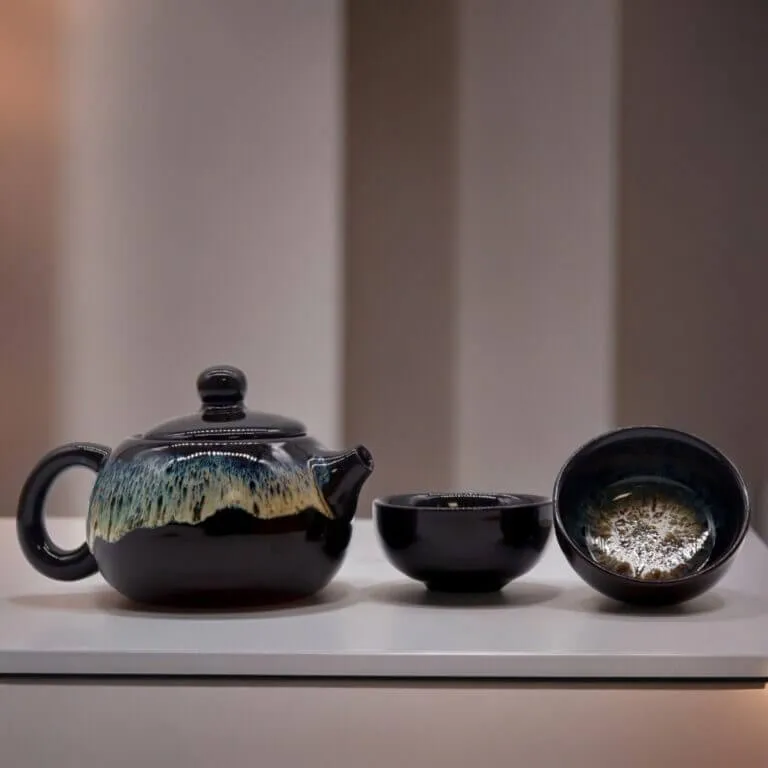 A black ceramic teapot with a textured glaze sits next to two matching teacups on a white surface against a softly blurred background.