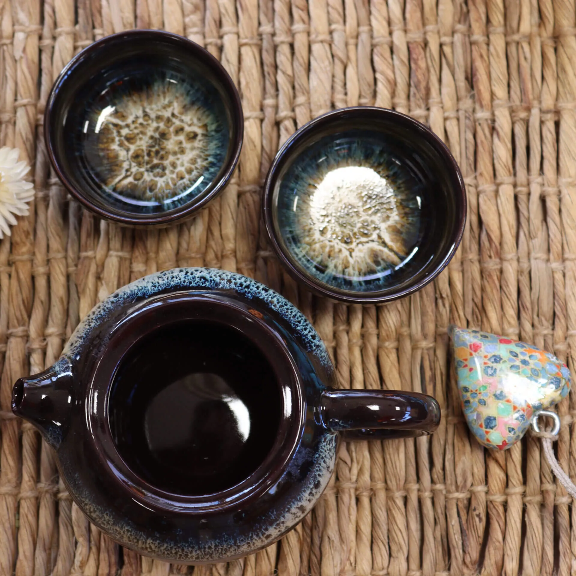 A dark ceramic teapot with a matching pair of cups and a floral-patterned ceramic heart on a woven mat.