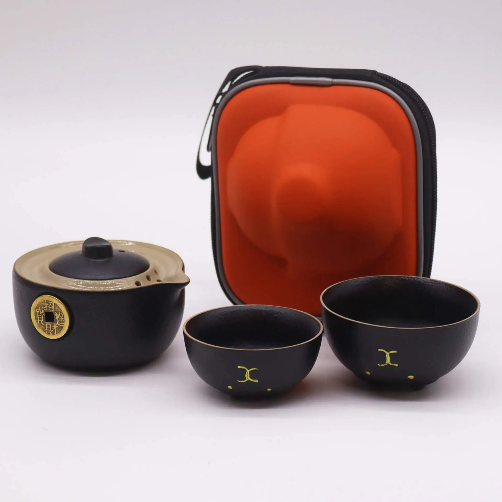 A black teapot with a lid, two matching teacups, and an orange protective carrying case are displayed on a white background.