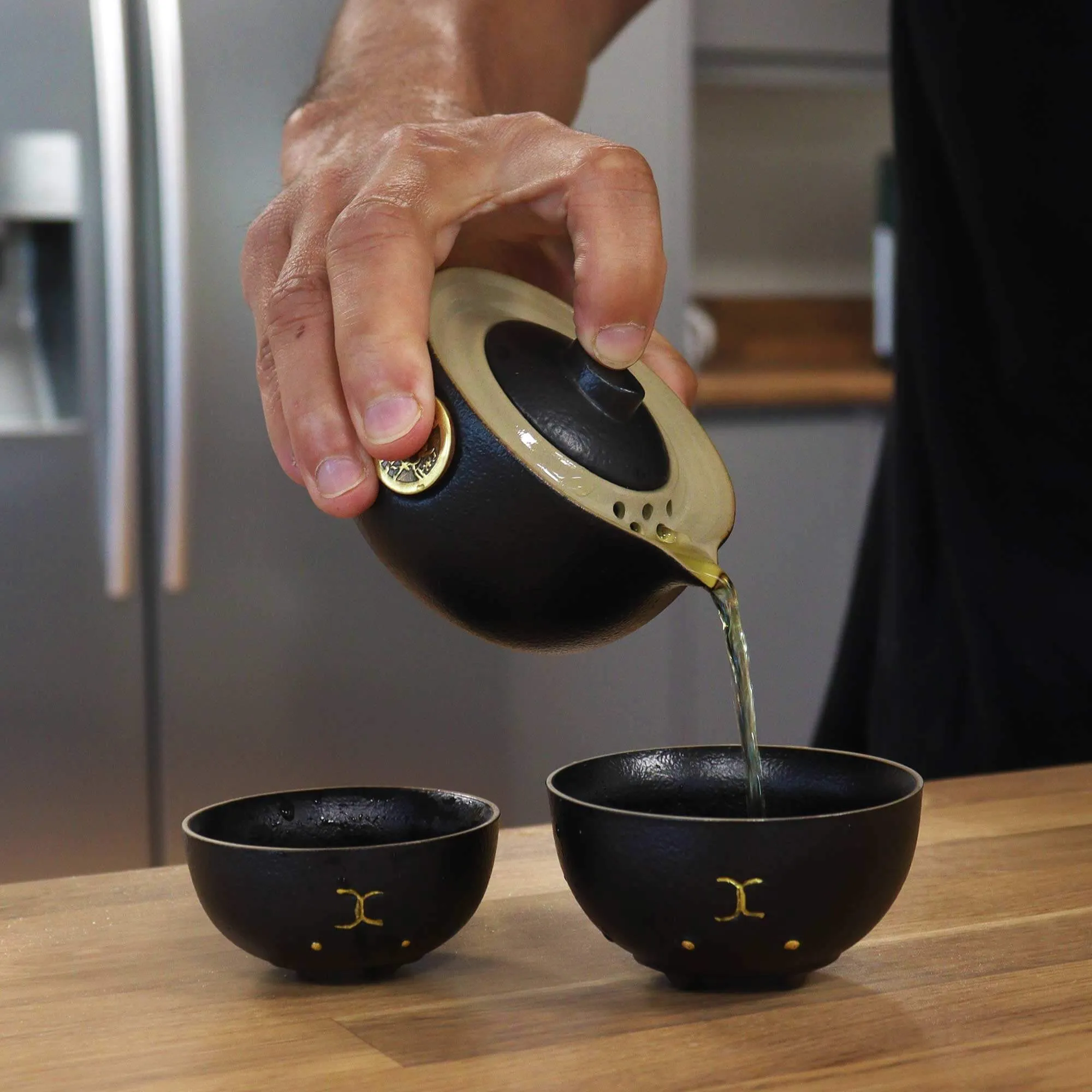 A hand pours tea from a small black teapot into one of two matching black teacups on a wooden countertop in a kitchen.