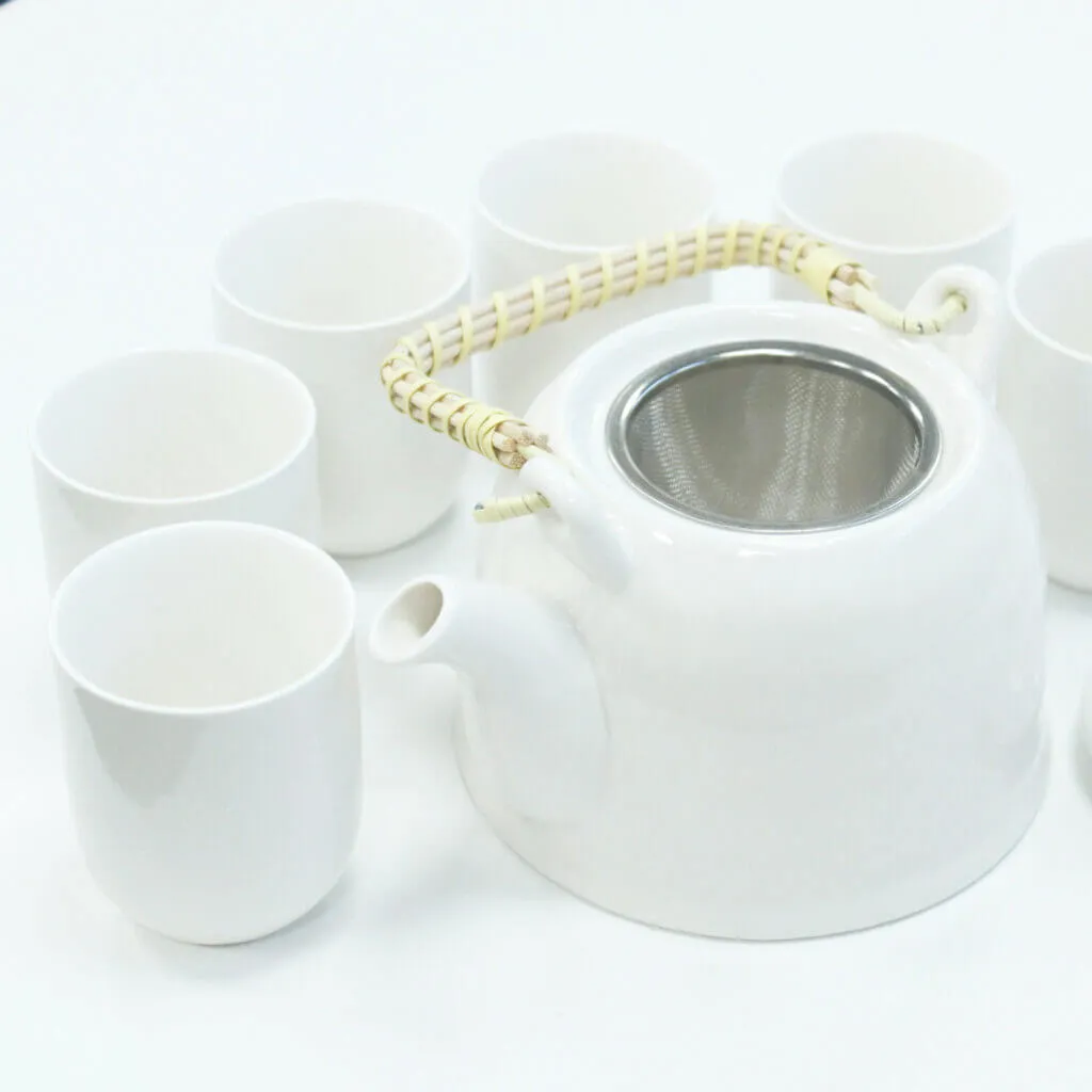 A white teapot with a woven handle and metal strainer is surrounded by six matching white cups on a white surface.