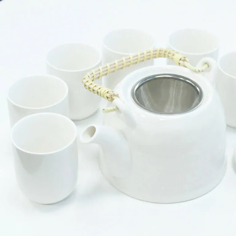 A white teapot with a woven handle and metal strainer is surrounded by six matching white cups on a white surface.