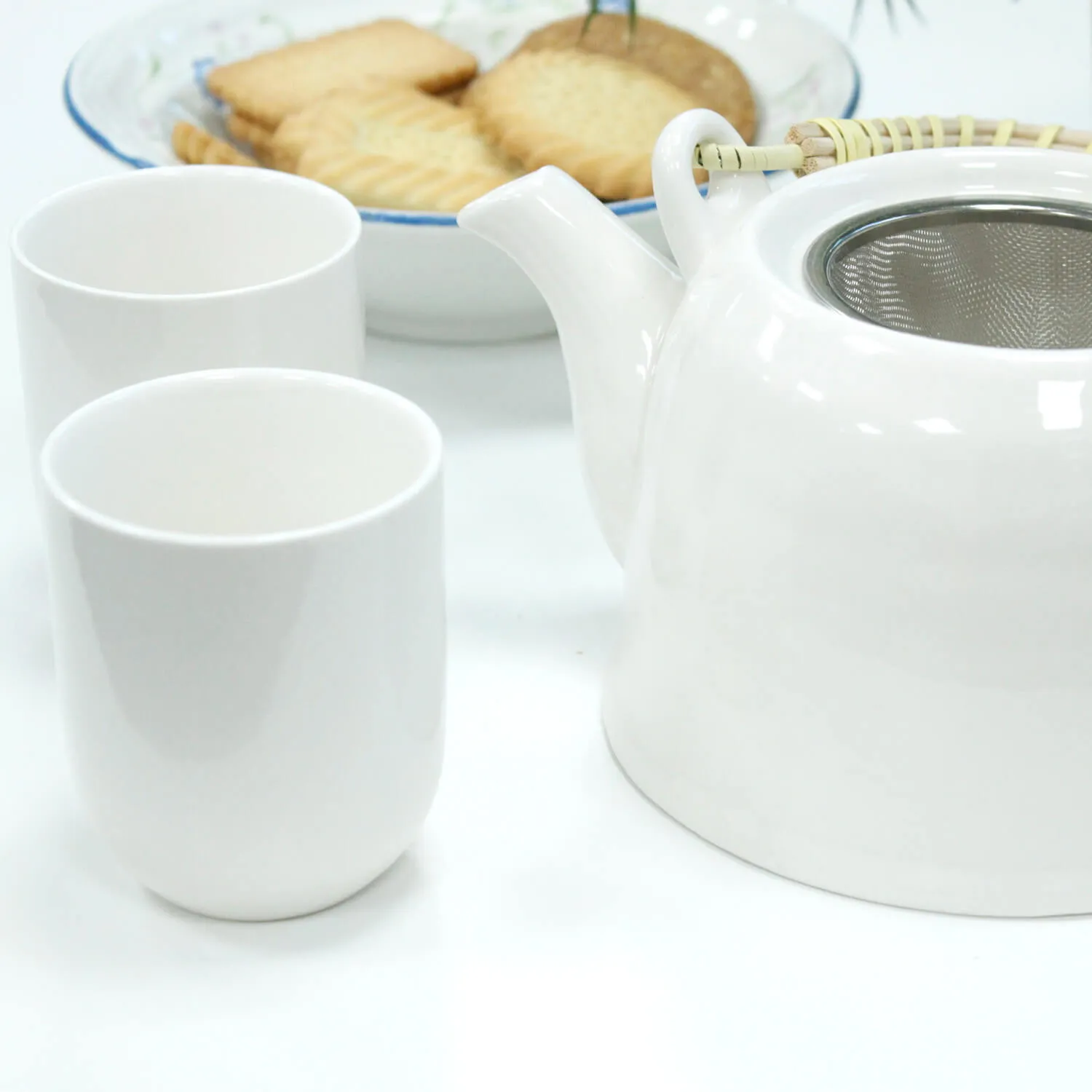 A white teapot, two white cups, and a plate of rectangular biscuits are arranged on a white surface.