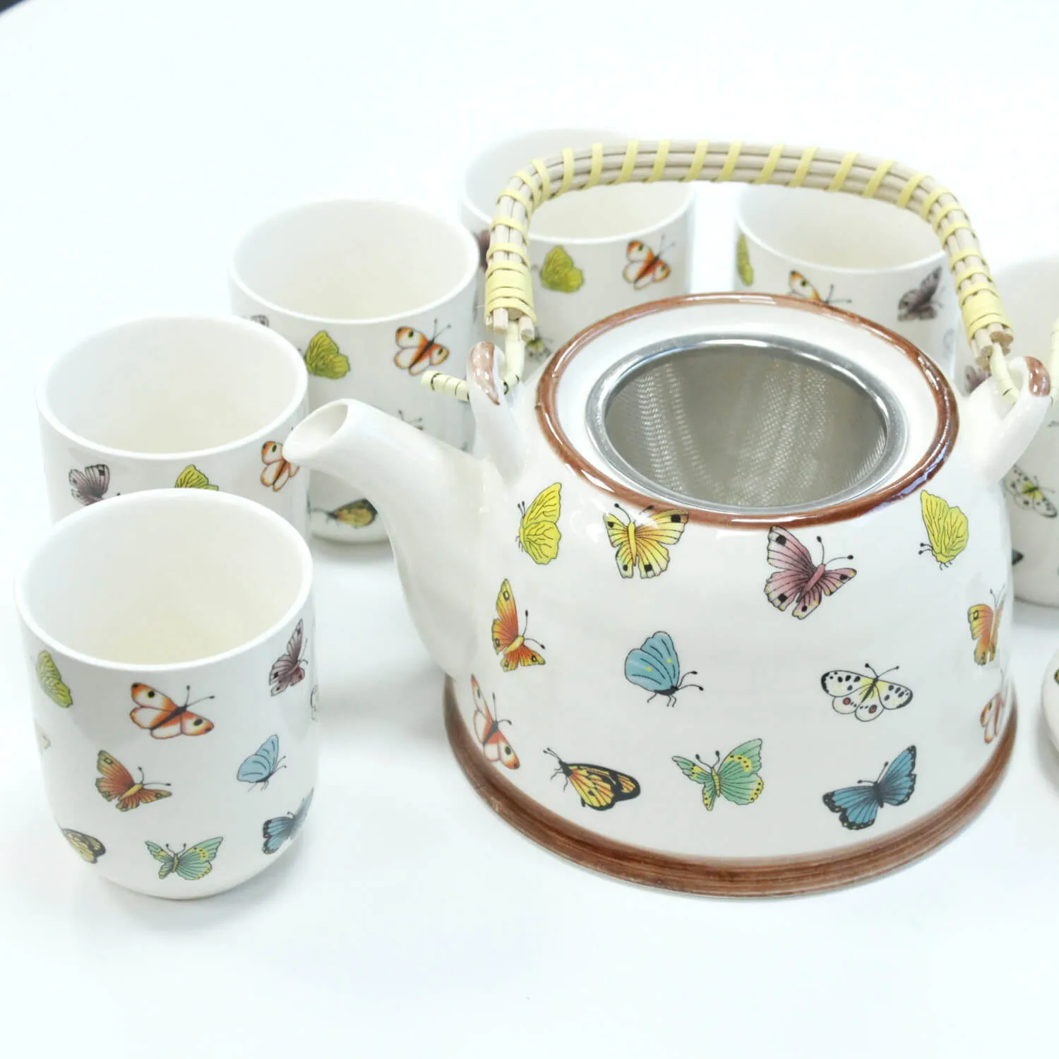 A ceramic teapot with a wicker handle and matching cups, all decorated with colorful butterfly illustrations, arranged on a white surface.