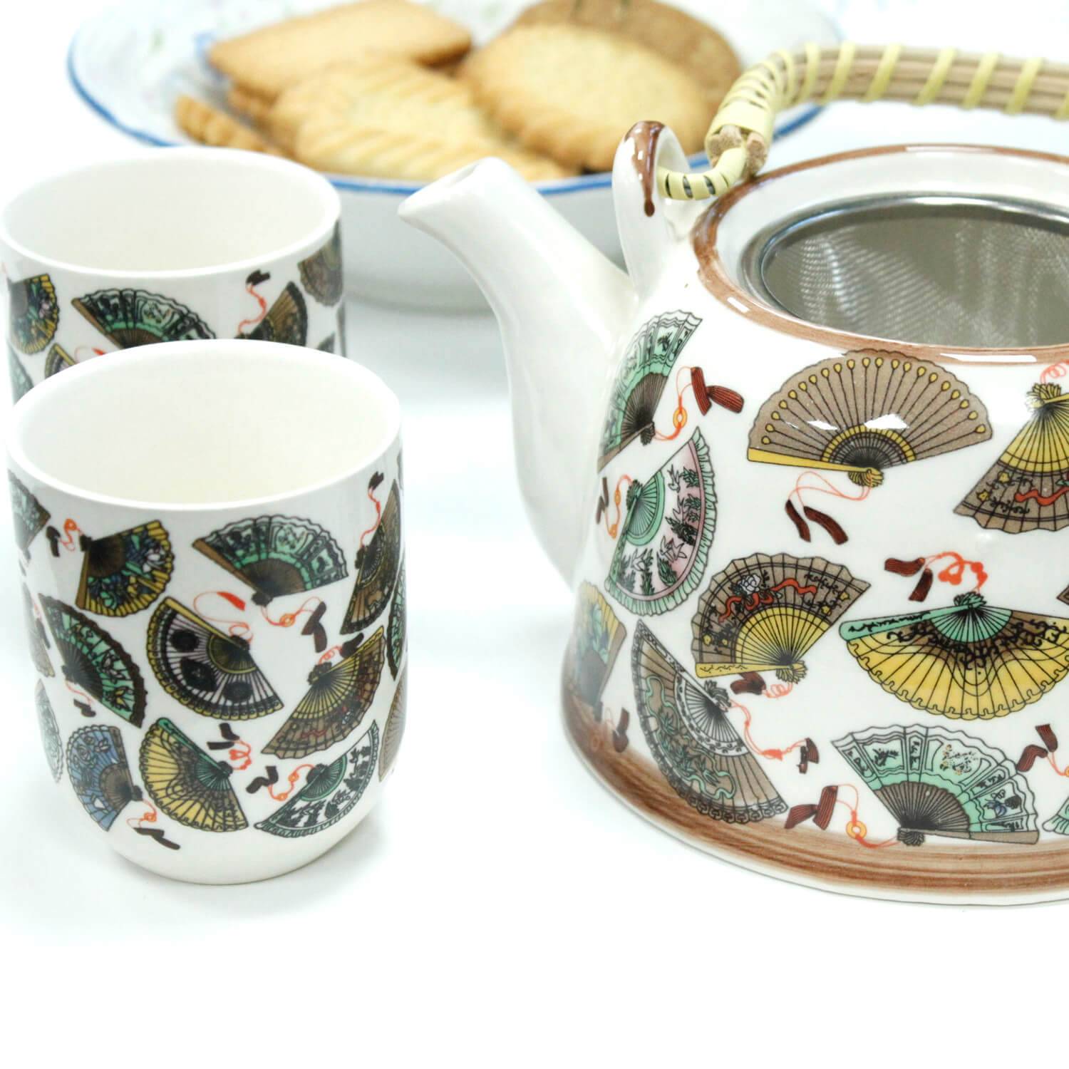 A ceramic teapot with matching cups decorated with fan patterns, next to a plate of rectangular cookies.