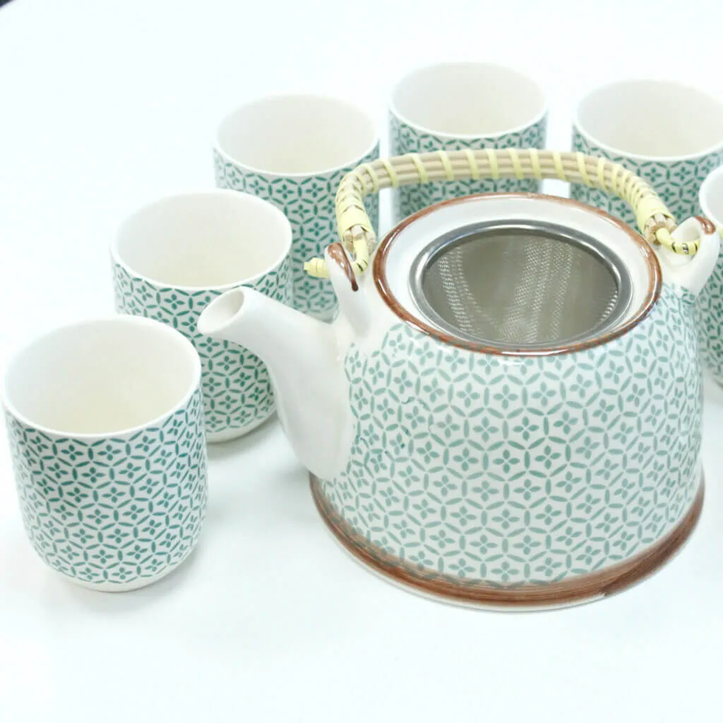 A patterned ceramic teapot with a wicker handle is surrounded by six matching teacups, all on a white surface.