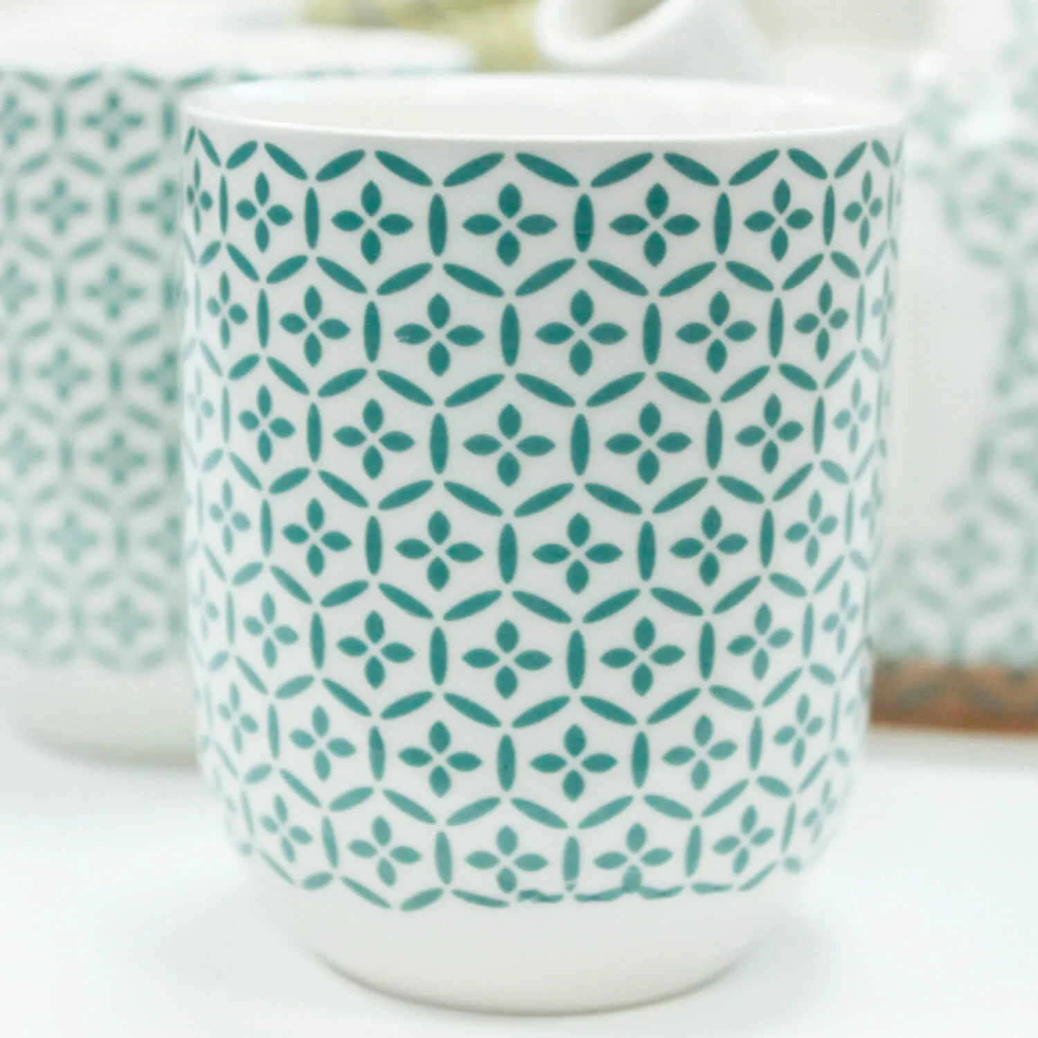 White ceramic cup with a teal geometric pattern consisting of repeating diamond and circle shapes. The cup is on a white surface, with another similar cup slightly blurred in the background.