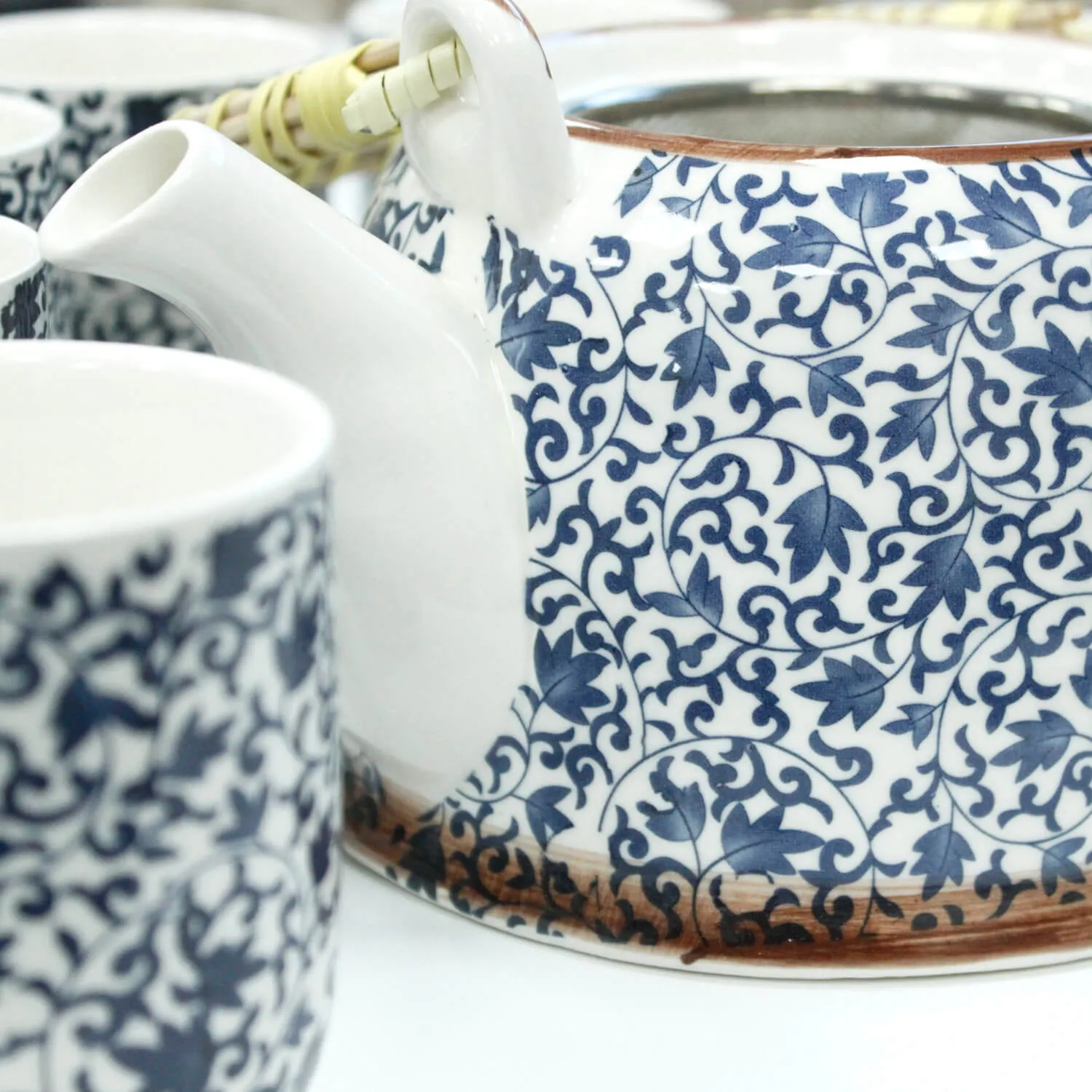 Close-up of a teapot and cups with a blue and white floral pattern. The teapot handle is partially visible. The pottery sits on a white surface.