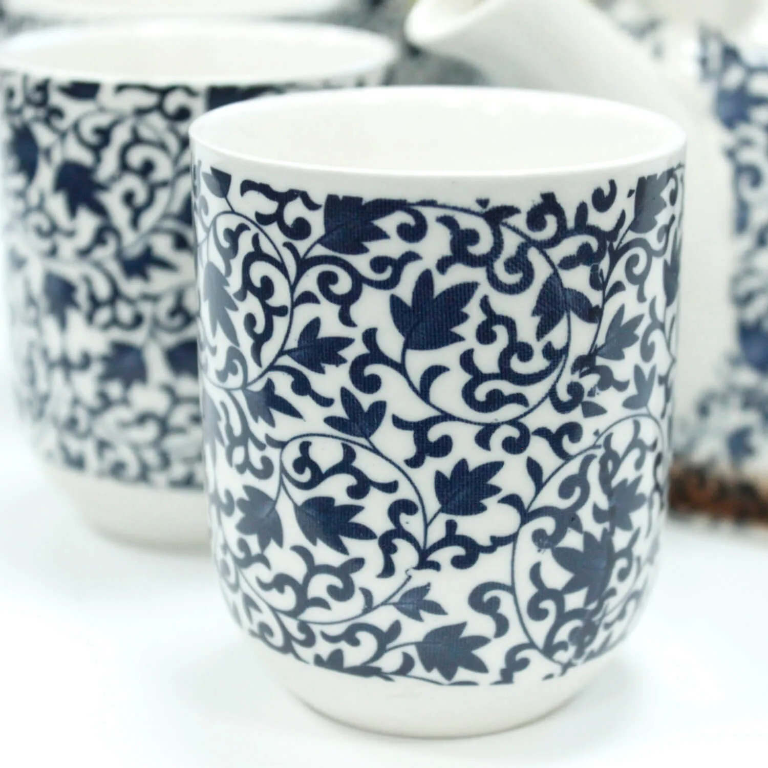 A white ceramic cup featuring an intricate blue floral vine pattern, with parts of matching cups and a teapot visible in the background.