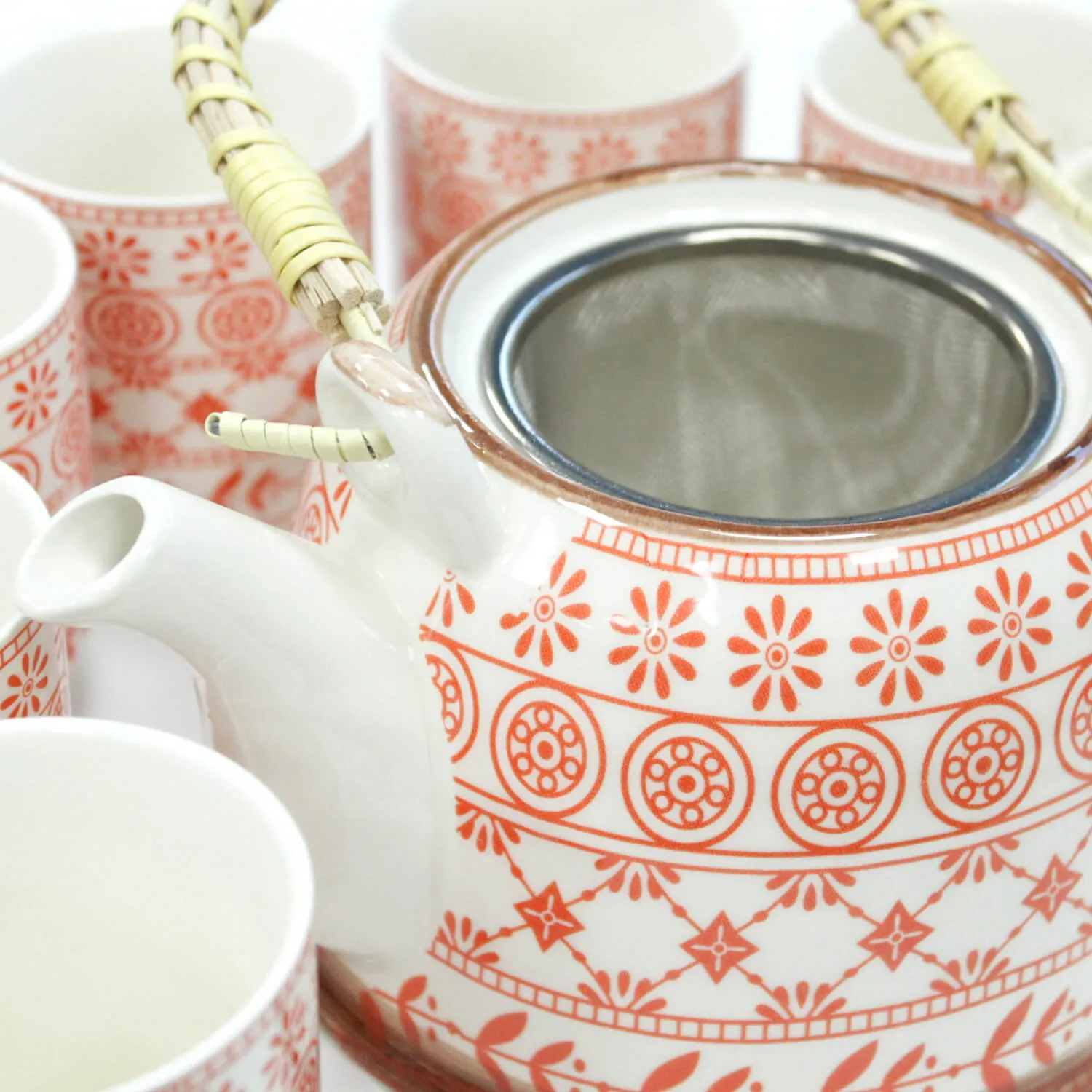 A white teapot with an orange floral pattern and woven handle surrounded by matching cups.