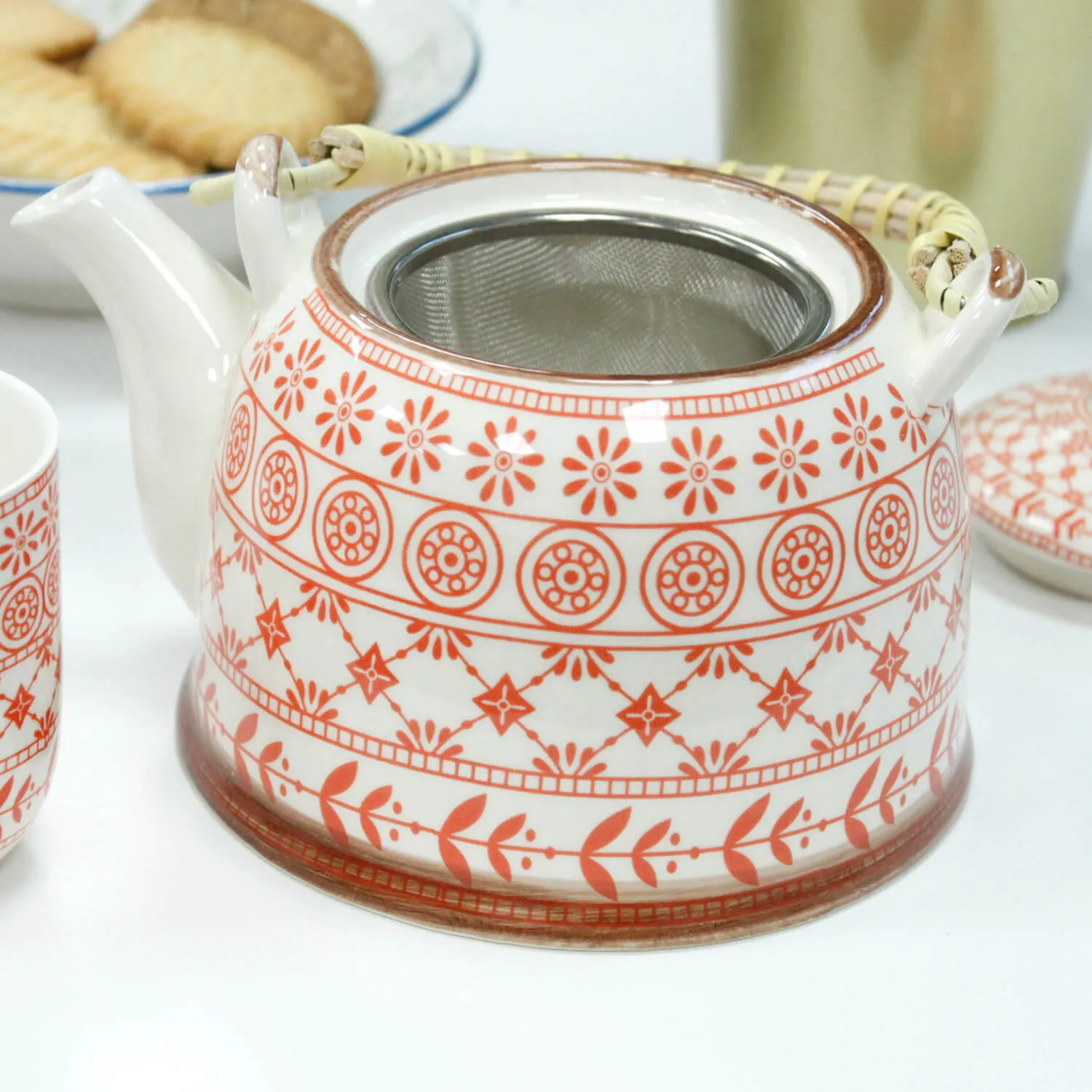 A ceramic teapot with a red geometric and floral pattern, a metal mesh infuser, and a bamboo-wrapped handle sits on a table near a teacup and plate of biscuits.
