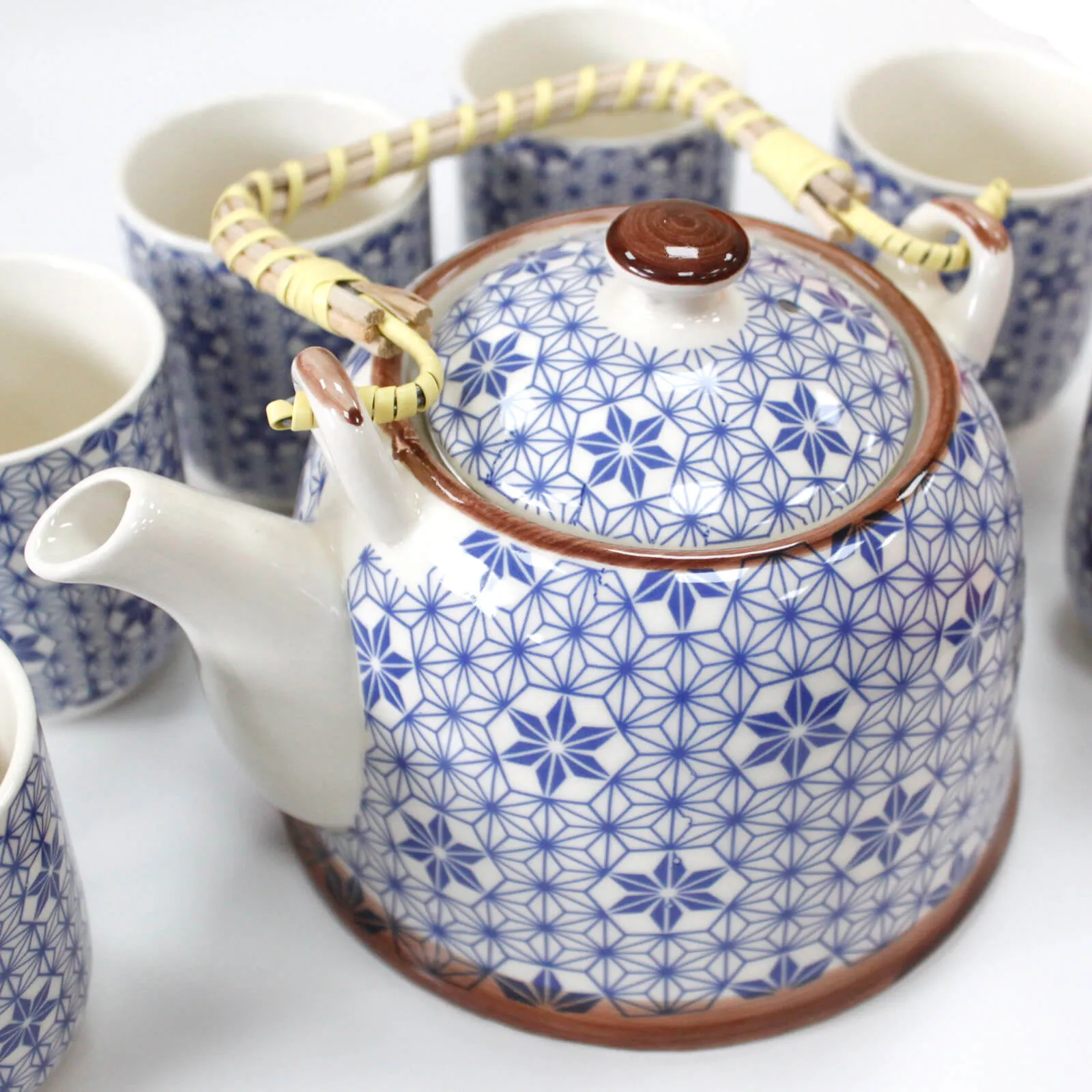 A ceramic teapot with a blue geometric pattern and woven handle is surrounded by matching teacups on a white surface.