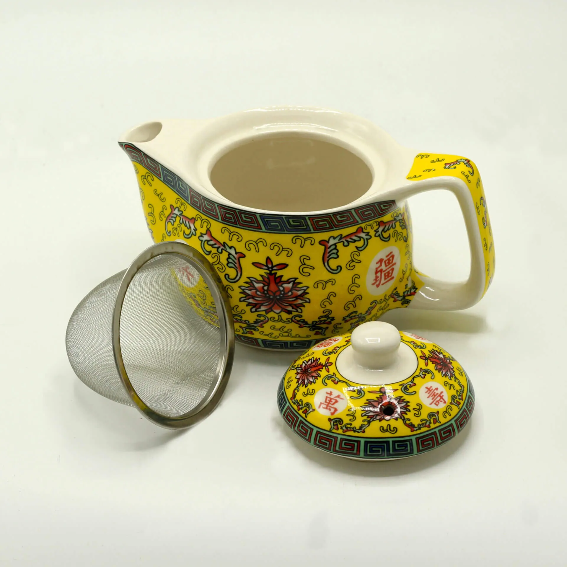 A yellow porcelain teapot with floral designs, its lid removed and placed to the side, alongside a metal mesh tea strainer.