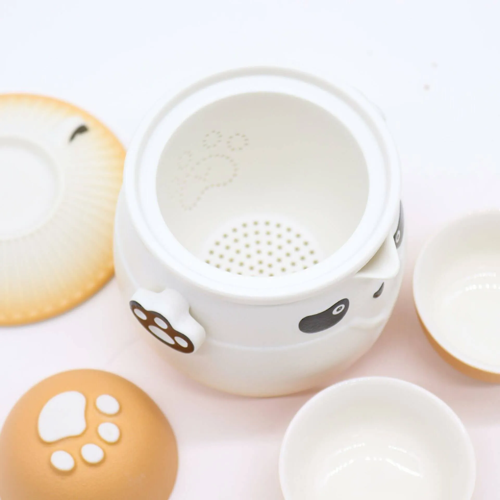 A white ceramic teapot with panda face and paw print designs, accompanied by a matching lid, two small teacups, and a saucer on a white surface.
