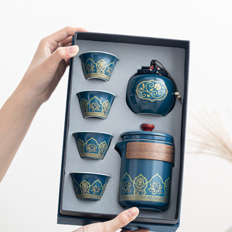 A person holds an open box containing a blue and gold tea set, including a teapot, four cups, and a small jar, all decorated with floral patterns.