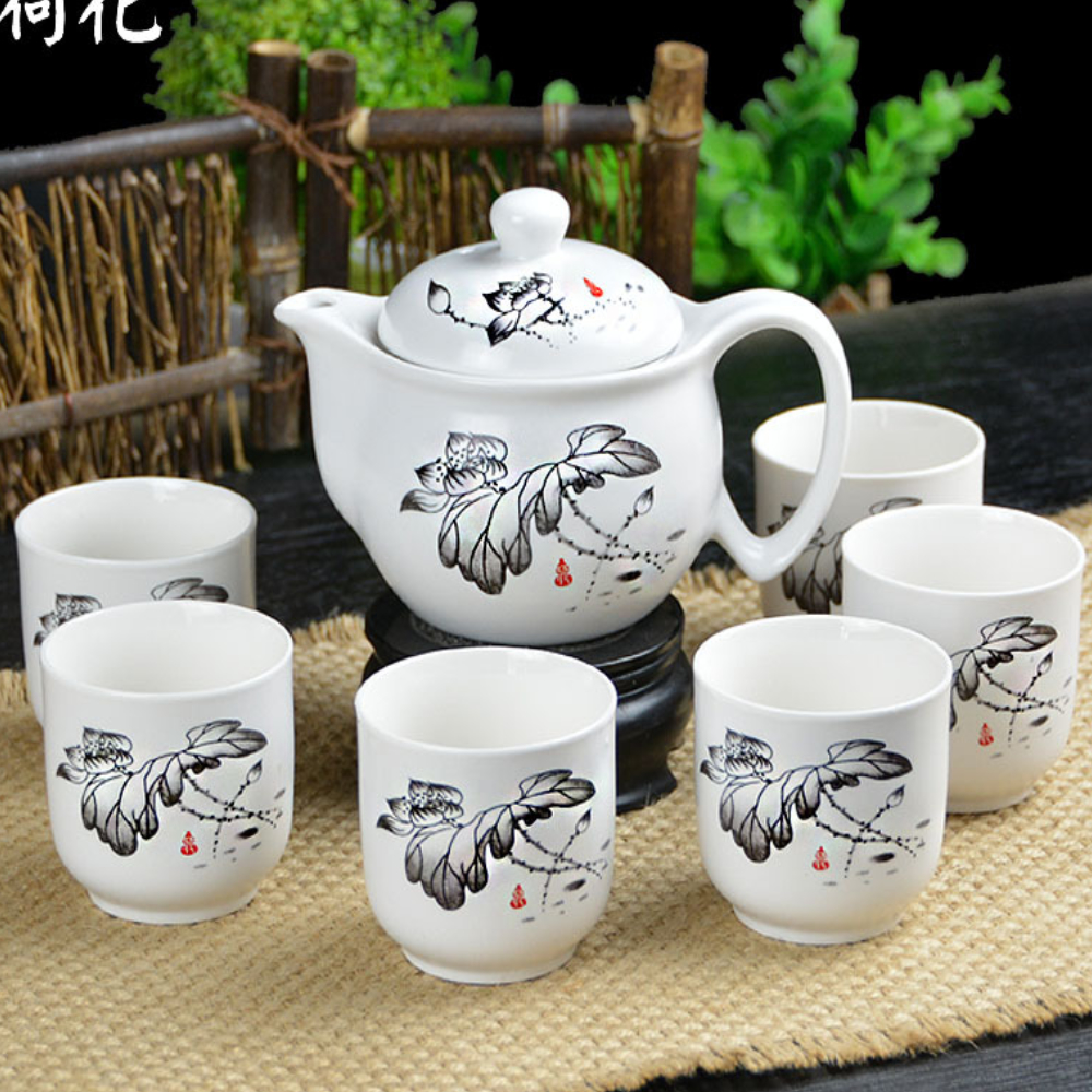 A white porcelain teapot with a lid and six matching cups, all decorated with black floral designs, are arranged on a woven mat.