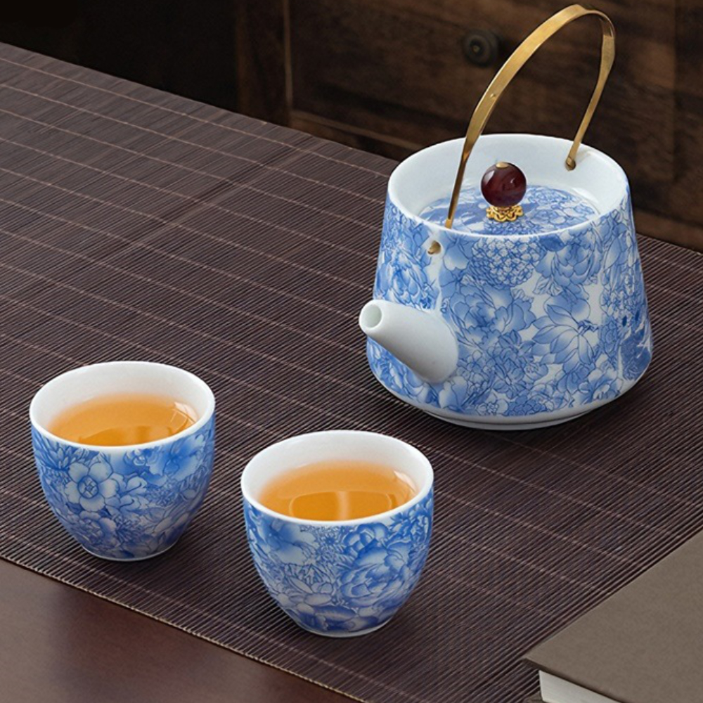 Blue and white floral porcelain teapot with a brass handle next to two matching teacups filled with tea, on a dark striped table mat.