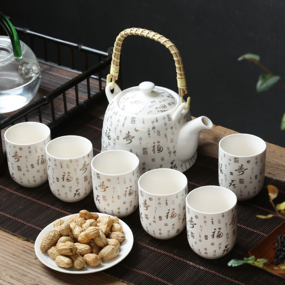A ceramic teapot with a bamboo handle and six matching cups with Chinese calligraphy are arranged on a mat, next to a plate of unshelled peanuts.