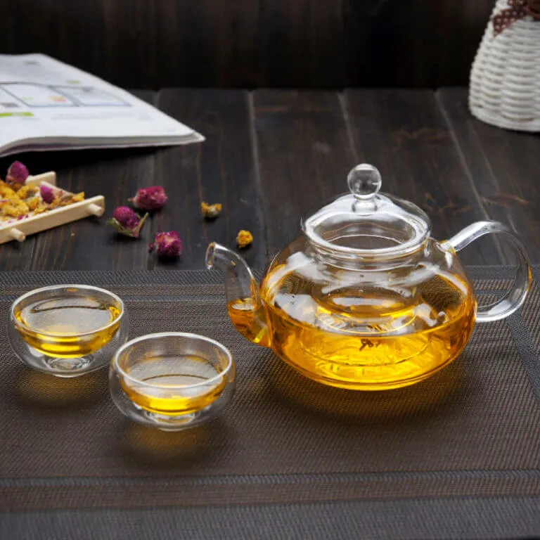 The Artisan Thick-Glass Teapot with premium borosilicate glass, filled with yellow tea, is accompanied by two small teacups on a woven placemat, surrounded by scattered rosebuds.