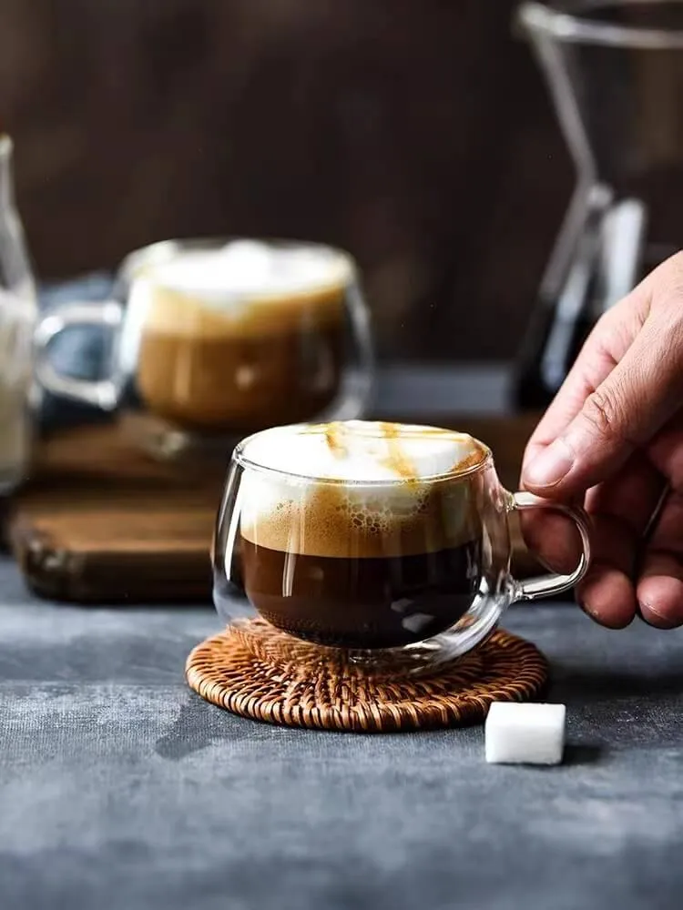A hand reaches for a Double-Wall Glass Cup | Floral Tea & Coffee Tumbler (150ml) filled with coffee and frothy milk on a woven coaster. A similar tumbler sits in the background, with a sugar cube nearby.
