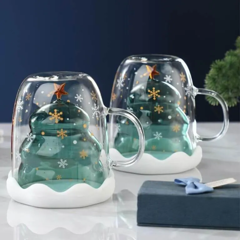 Two glass mugs with inverted Christmas tree designs inside, decorated with snowflakes and a star on top, placed on a marble surface next to a blue gift box.