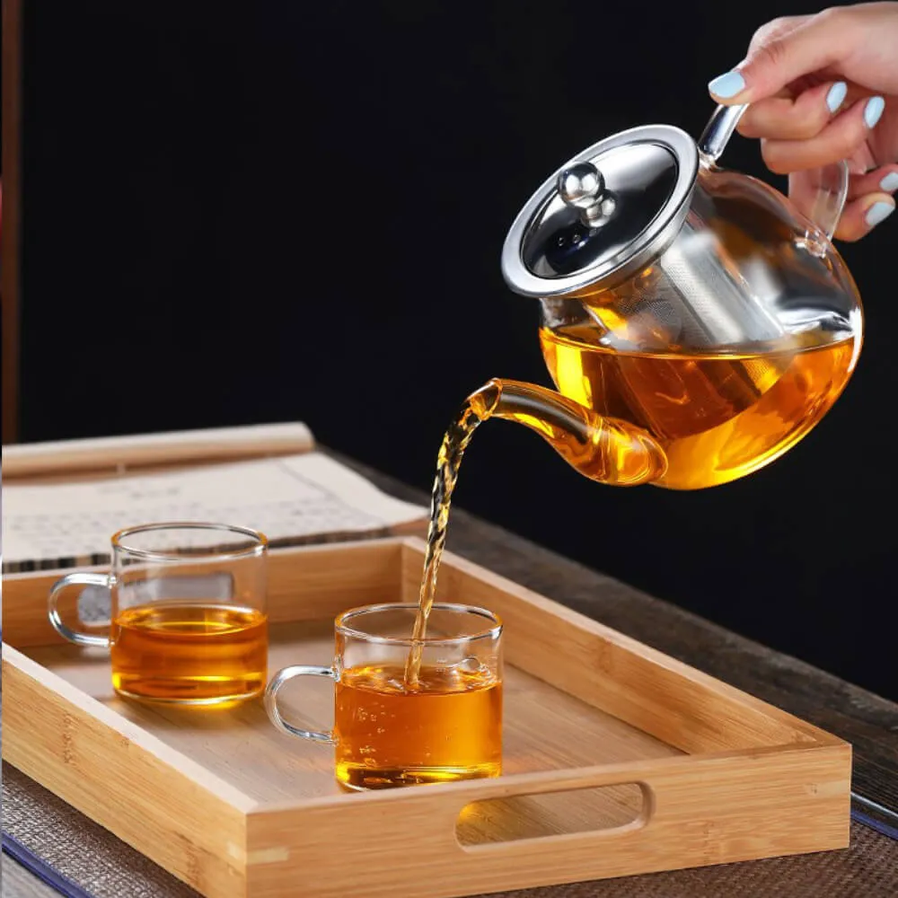 A hand pours tea from the Elegant Glass Teapot with Stainless Steel Infuser (800ml) into a cup, while another cup rests on a wooden tray next to a closed book.