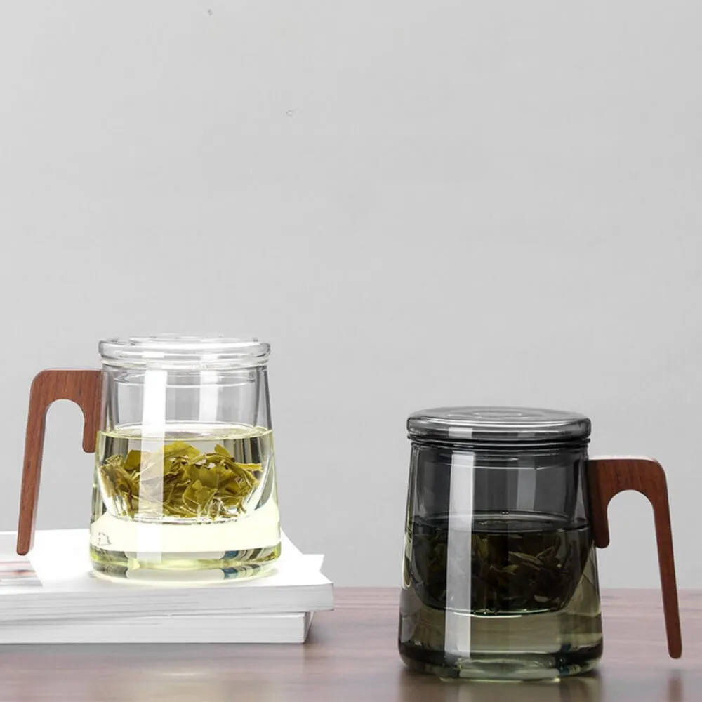 Two Modern Glass & Walnut Tea Infuser Cups in smoke gray sit elegantly on wood—one atop books and one directly on the table. Each showcases brewed tea and leaves, embodying the office tea master's refined taste.