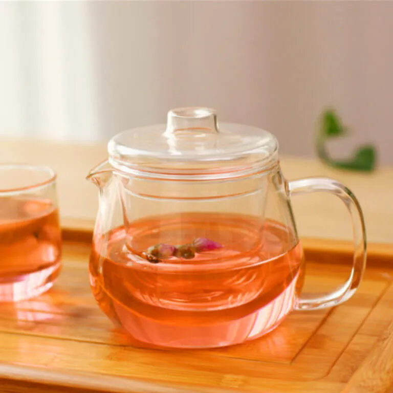 Next to a glass cup on a wooden tray, the Hand-Blown Penguin Glass Teapot with Flower Filter sits elegantly with pink herbal tea and floating ingredients, enhancing your tea experience both aesthetically and in flavor.