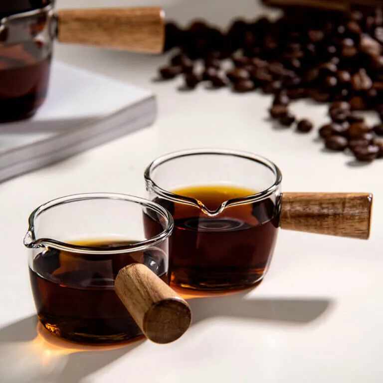Two Italian-Style Glass Milk Pitchers with wooden handles are filled with coffee on a white surface. Coffee beans and a closed book in the background create a serene scene.