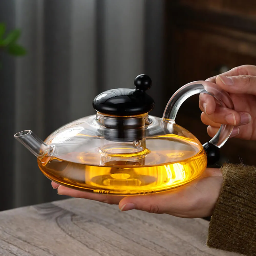 A person cradles the Nordic Lucky Rat Tail Glass Teapot, showcasing a sleek black lid and handle, its amber tea glowing warmly. The elegant design features a built-in tea infuser and rests on wood, set against a softly blurred background.