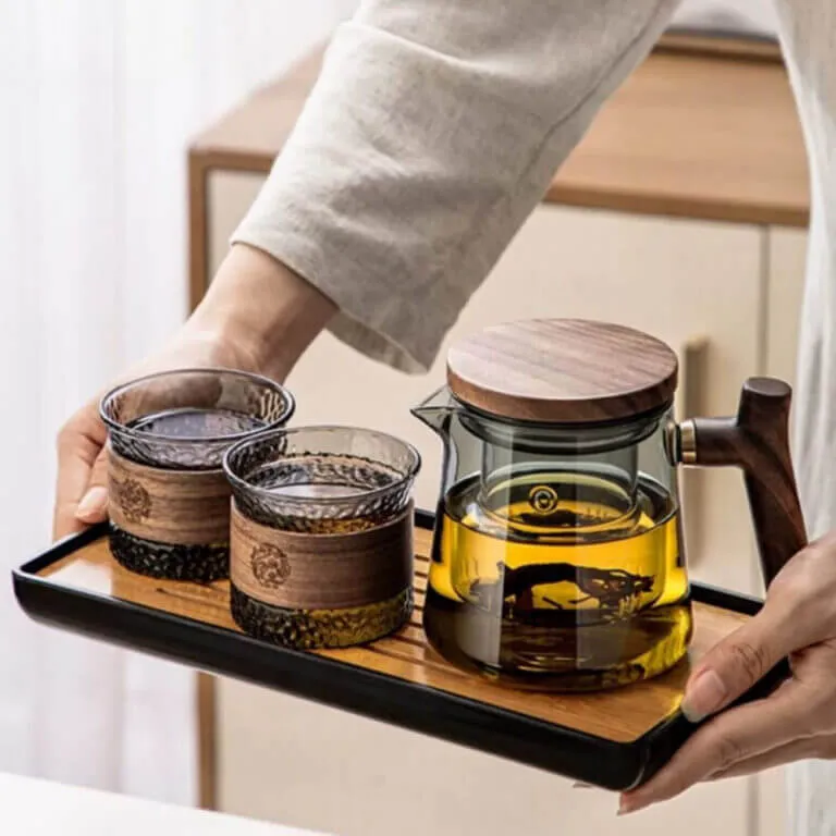 Someone showcases a Personal Glass Tea Master | Single-Serve Office Brewing Set, with a glass teapot containing yellow liquid and two glasses, all featuring wooden accents.