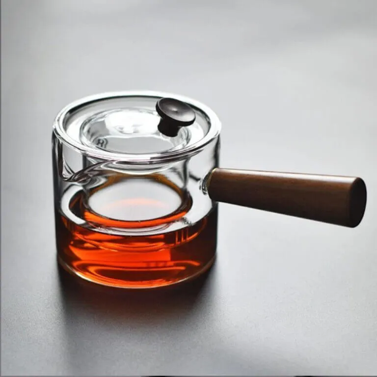 The Japanese-Style Glass Teapot with Wooden Side Handle in a Modern Minimalist Design shows amber liquid on a gray surface.