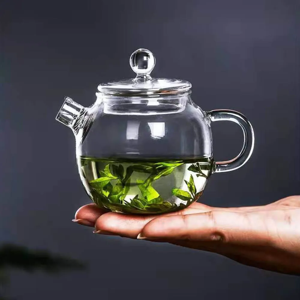 A Petite Glass Bubble Teapot with Lid, brimming with green tea leaves, is elegantly held in a person's hand against a dark backdrop.