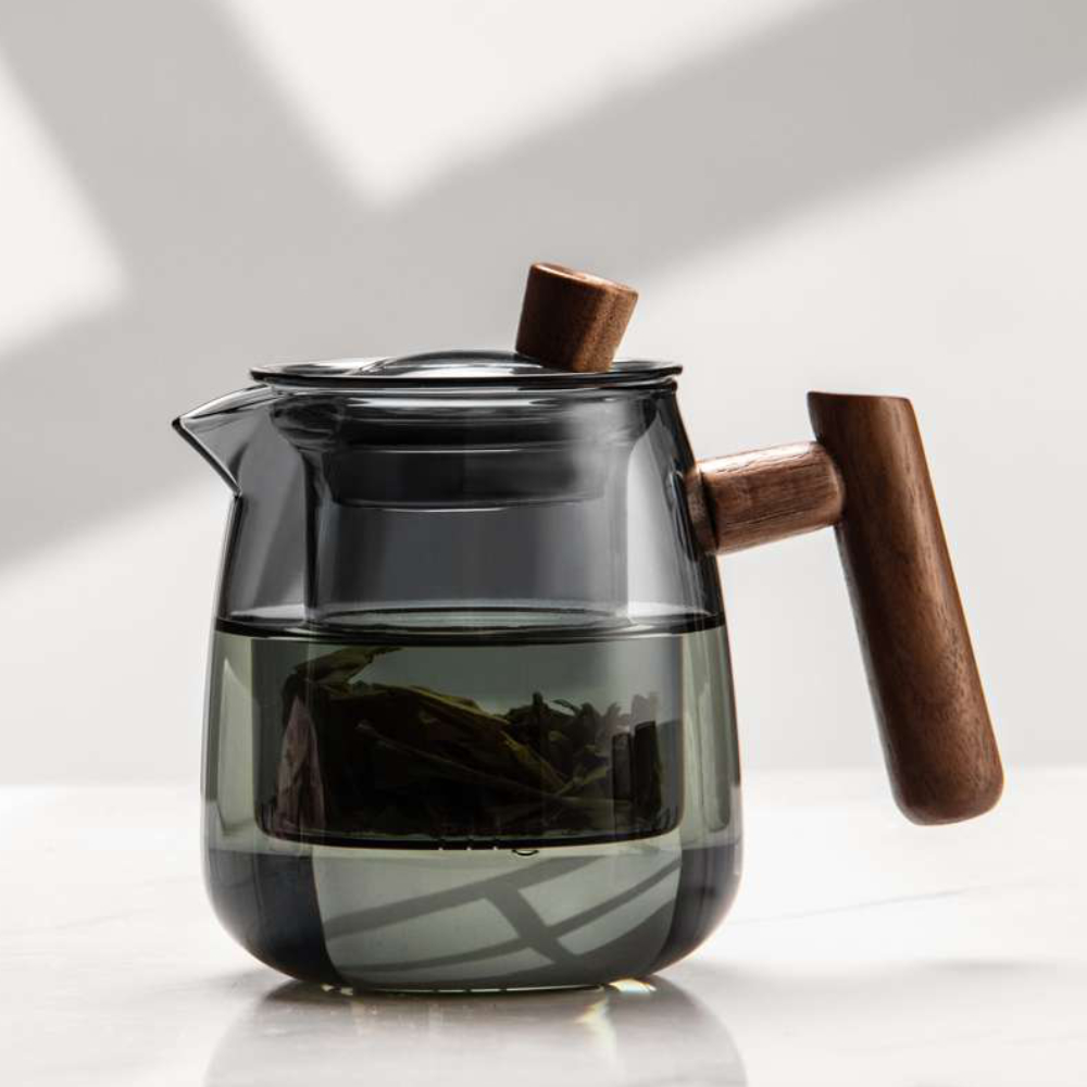 The Mountain View Glass Teapot, featuring a Japanese-style side handle with a wooden accent, rests on a white surface filled with water and loose tea leaves.