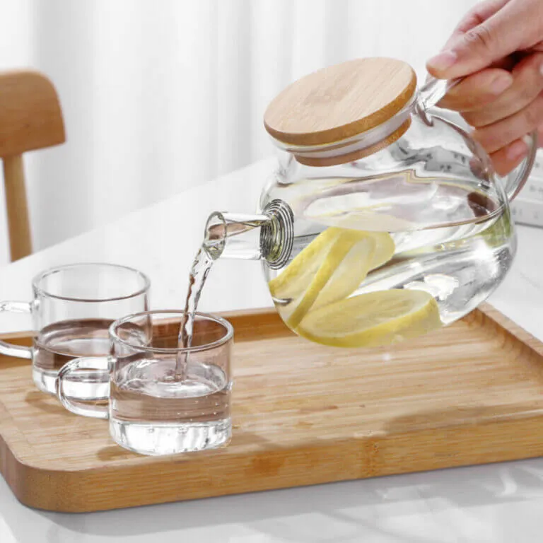 A Nordic Glass Teapot with a Bamboo Lid & Steel Spout gracefully pours lemon water into a glass on a wooden tray, next to an empty glass.