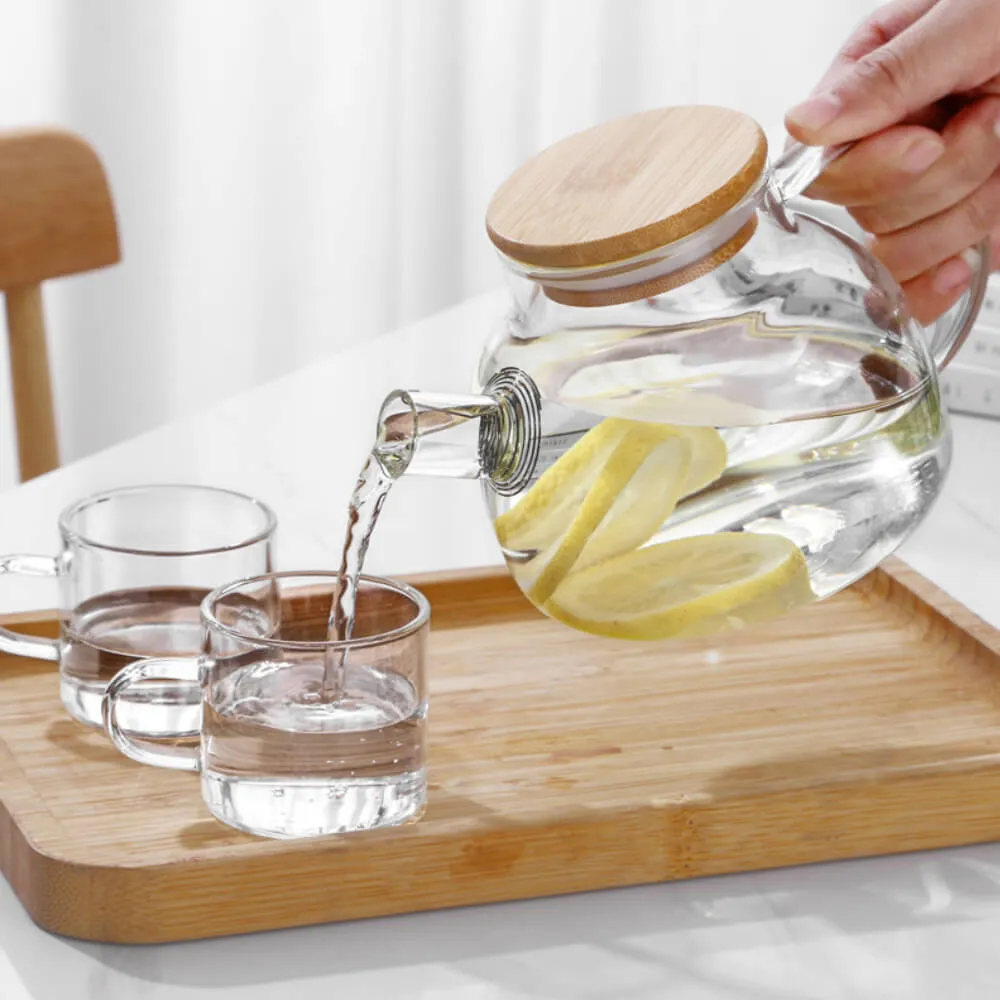 A Nordic Glass Teapot with a Bamboo Lid & Steel Spout gracefully pours lemon water into a glass on a wooden tray, next to an empty glass.